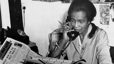 Claudia Jones, a Black woman, is answering a phone with a pen in hand, near a typewriter and a copy of the West Indian Gazette. Headline reads - What Next for the West Indies?’