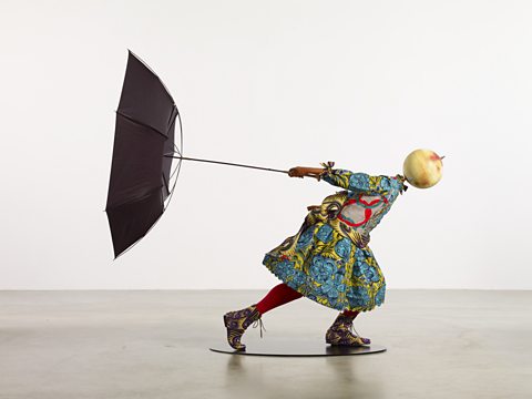 A sculpture by Yinka Shonibare. A figure holds an umbrella out behind them. The figure is made of lots of different patterned fabrics.
