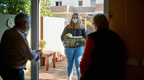 A young woman delivering a box of groceries to an elderly couple through a screen door while wearing a mask and gloves.