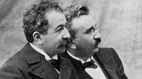 Auguste Lumiere, an older white man with a moustache, poses with Louis Lumiere, who also has a moustache and glasses.