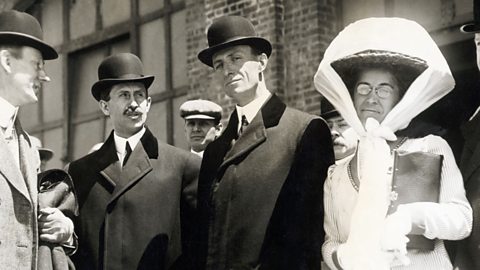 Wilbur and Orville Wright, both in coats and bowler hats, and Katharine Wright stands with a sash tied around her hat.