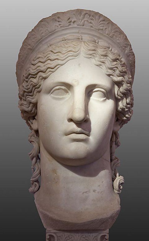 A white marble statue of the head of a woman on a small plinth - details show long wavy hair held back at the ears and on her head by a band