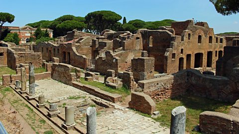 In front of a blue sky and some trees are the brick ruins of insulae dwellings, many walls and the roofs are missing. Some stubs of stone pillars can be seen also.