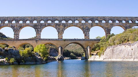 An aqueduct built from stone crosses over a body of water to the ground at either side. It has three levels of arches - the uppermost contains smaller arches.