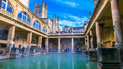 A large pool of water with steam rising from it to the open air above is surrounded by ornate stone architecture, walkways and pillars.