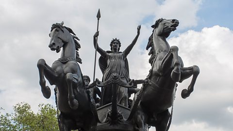 A bronze statue of Boudica - spear in hand with her arms raised. She stands on her chariot, which is led by two horses rearing up on their hind legs.