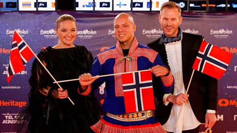 Norway's Keiino pose for a picture during the Red Carpet ceremony. The middle male figure is wearing a traditional Sami outfit. All three are holding Norwegian flags.