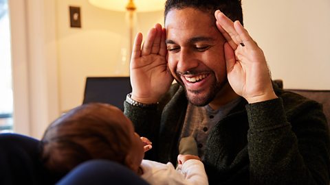 Smiling young father playing peekaboo with his adorable baby boy lying on his lap on a sofa at home.