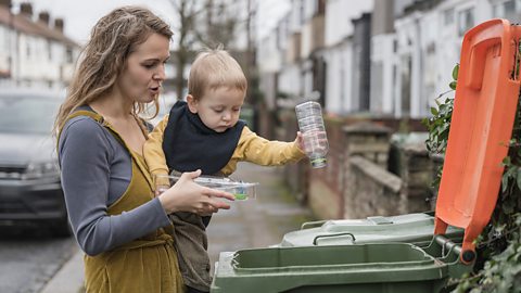 Partial side view of long-haired woman in casual clothing holding 2-year-old son in her arms as he puts plastic containers in recycling bin.