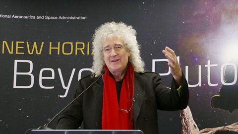 Brian May, guitarist of rock band Queen, talks using a microphone standing in front of NASA's New Horizons mission signs.