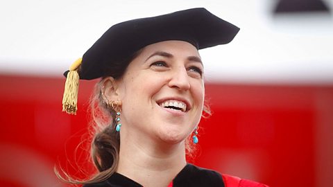 Actress Mayim Bialik wearing a black university gown and hat, smiling.