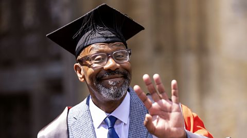 Sir Lenny Henry waving at the camera, wearing a university hat and gown over a grey suit and tie.