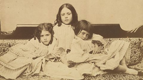 Edith, Lorina and Alice Liddell, three young girls in gowns sat together on a sofa.