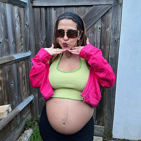 Suzie Bajos in a green crop top and pink jacket, showing her baby bump and making a kissing face. She is standing in a back garden in front of a face.