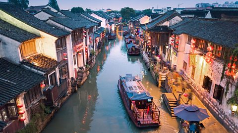 A wooden canal boat travels down a canal at dusk. Along the canal are traditional Chinese buildings with soft lighting.