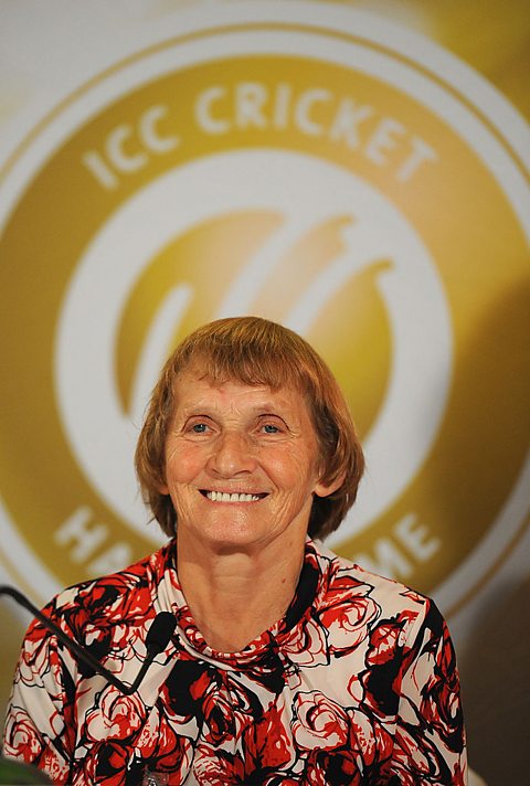 A photo of a smiling older woman with a brown bob and wearing a red and white floral shirt. She sits in front of a gold and white background.