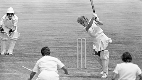 A black and white photo of a woman with short hair and wearing white clothing, batting in a cricket match, with three fielders visible.