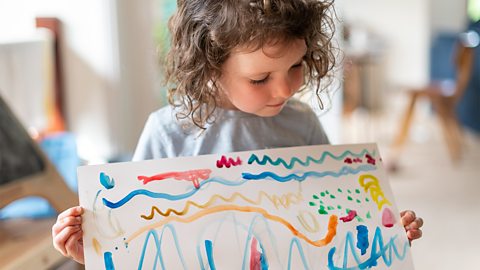 A little girl holding up a sheet of paper with lots of coloured paint squiggles.