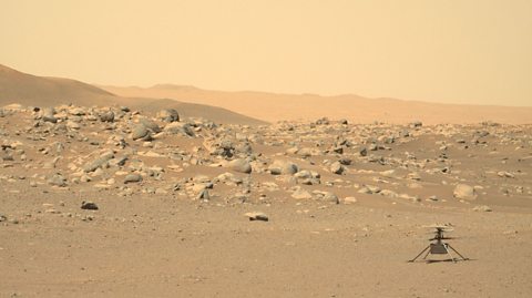 Photo from Perseverence Mars Rover of Ingenuity the mini helicopter - the sky and ground are dusty and there is lots of rocks and boulders around.