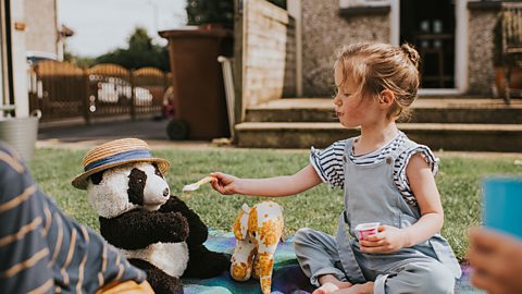 Cute young siblings sit on a rug outside on a warm day, along with several soft toys, enjoying a teddy bear's picnic lunch. Main focus is on realistic panda bear soft toy, wearing a sun hat and the young girl spoon feeding him.