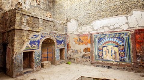 A photo of two walls of an ancient ruin building. Brightly coloured mosaics, with lots of shades of blue, feature on both walls. On the right mosaic, two figures can be seen in the mosaic.