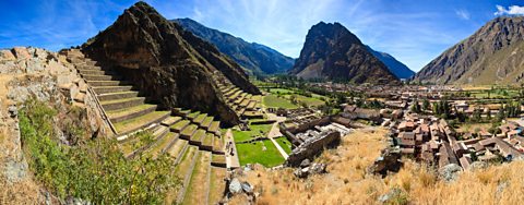 A panoramic landscape photo of the ruins of Ollantaytambo, with steep stone terraces cut into the hills on the left and stone buildings on the lower lying ground on the right