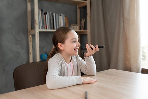 A young girl in a pink shirt with a grey cardigan is sitting at a wooden table with a bookshelf behind her. She is smiling and speaking into a phone.