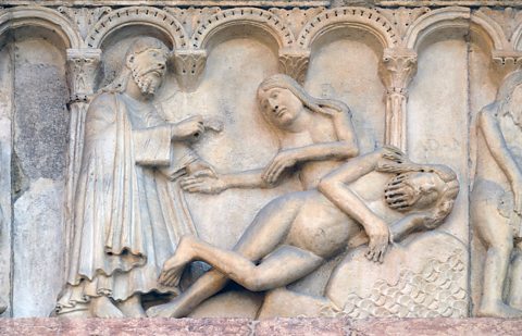 A stone carving of a man in a robe holding the hand of a female who is appearing from inside a sleeping man's body.