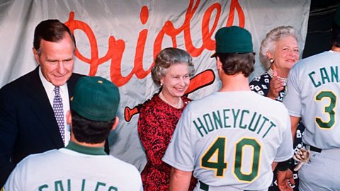 A line of male baseball players wearing white shirts and green caps shakes the hands of President Bush (wearing a suit), Queen Elizabeth II (wearing a red dress) and Barbara Bush (wearing a blue dress)