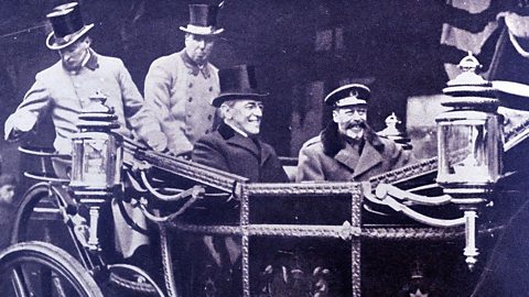 A black and white photograph of President Wilson and King George V sat in an open-tipped horse drawn carriage. Both are wearing hats and heavy coats. 