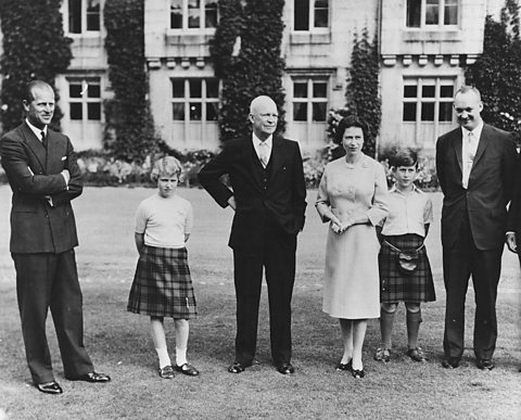 A black and white photograph of President Eisenhower with the Royal Family in front of Balmoral Castle. The men wear suits. whilst the children wear kilts. 