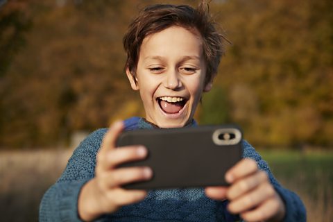 A boy in a blue jumper is smiling enthusiastically as he looks into a phone screen.