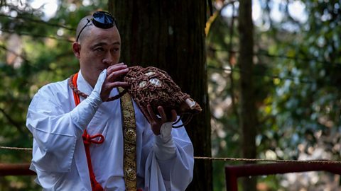 A bald Japanese man wearing a small black hat and long white robe, with a gold sash, plays music on a large conch shell wrapped in rope