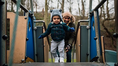 A dad standing behind his son on the play equipment at the park.