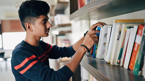 A teen boy looks at books on a shelf in a college-like setting