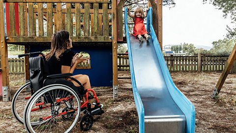 Smiling mother in a wheelchair playing with her little daughter on a slide in a playground. Children's games concept.