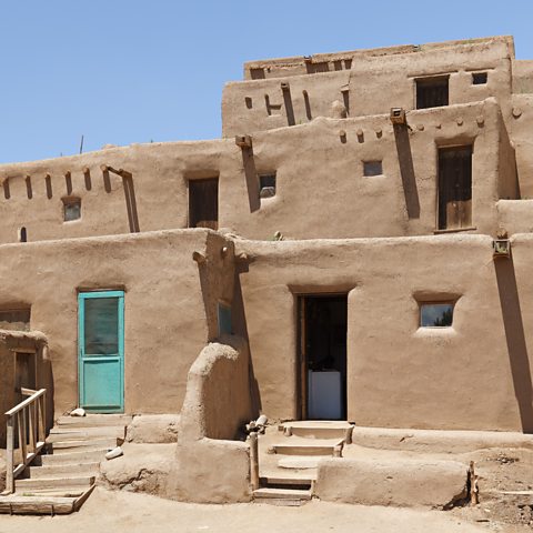 Photograph of light brown mud built adobe houses with small square windows against a blue sky. One of the houses has a turquoise door.