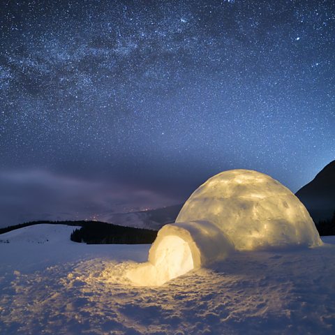 A photo of an igloo lit up from inside. It is night time and the sky is full of stars. The igloo is surrounded by snow.