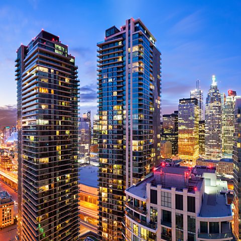 Photo showing two tall skyscrapers with other tall buildings in the background. It is evening and some of the lights in the buildings are on.