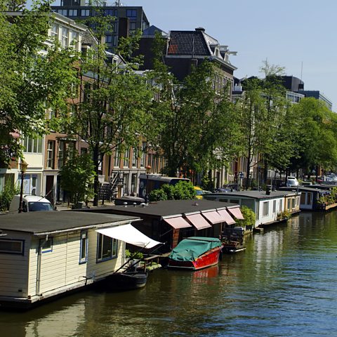 Photograph showing houseboats along a river in Amsterdam. Some have awnings out over their windows. There are a row of trees and a row of terraced houses behind them.