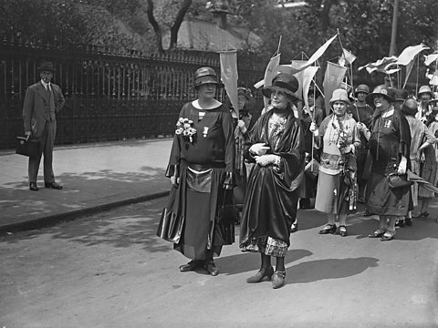 Two women standing in the the road in front of a crowd of women each carrying a banner 