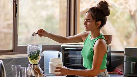 Woman stands in kitchen making a protein shake, seen putting a scoop of protein powder into a blender 