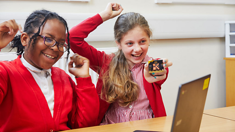 Two female primary school pupils hold up a micro:bit in front of a laptop