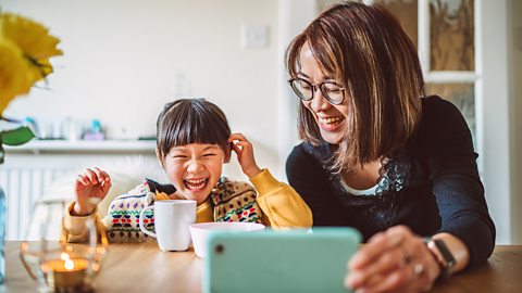 Young pretty Mom & her lovely little daughter having video call joyfully on smartphone while enjoying snacks on a dining table at home.