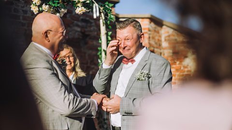 Smiling groom wiping his tears with finger while holding hand of partner at their wedding