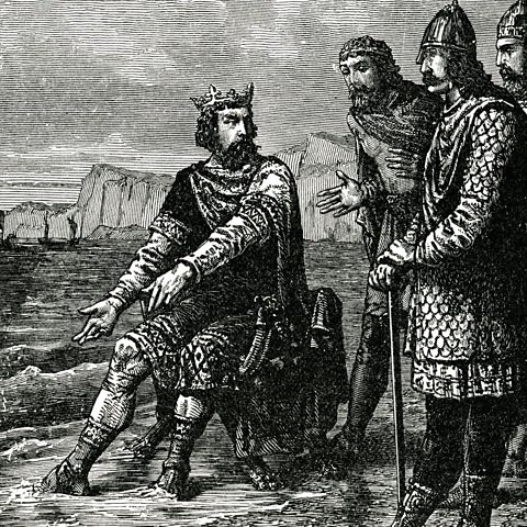 A back and white engraving of King Canute sitting on the seashore with water lapping his feet, turned around to look at some of his knights.
