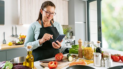 A woman stands at the kitchen counter with ingredients in front of her (jar of pasta, pepper, courgette, tomatoes, carrots and lettuce)