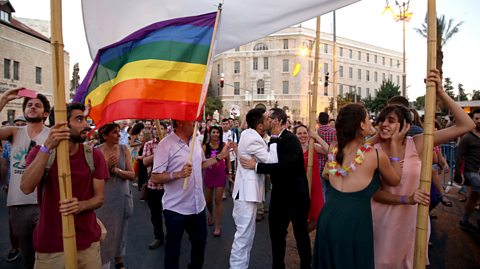 A parade with someone hold up a Pride flag next to a gay couple kissing under a huppah.