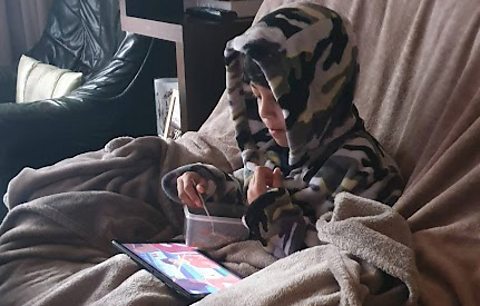 Little boy sits on sofa eating a snack as he uses his screen time to watch a tv show on his tablet