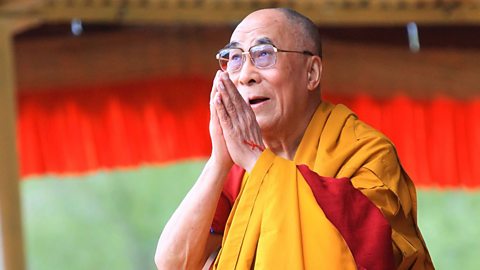 A man (the Dalai Lama) holds up two hands in a prayer position whilst looking forwards.